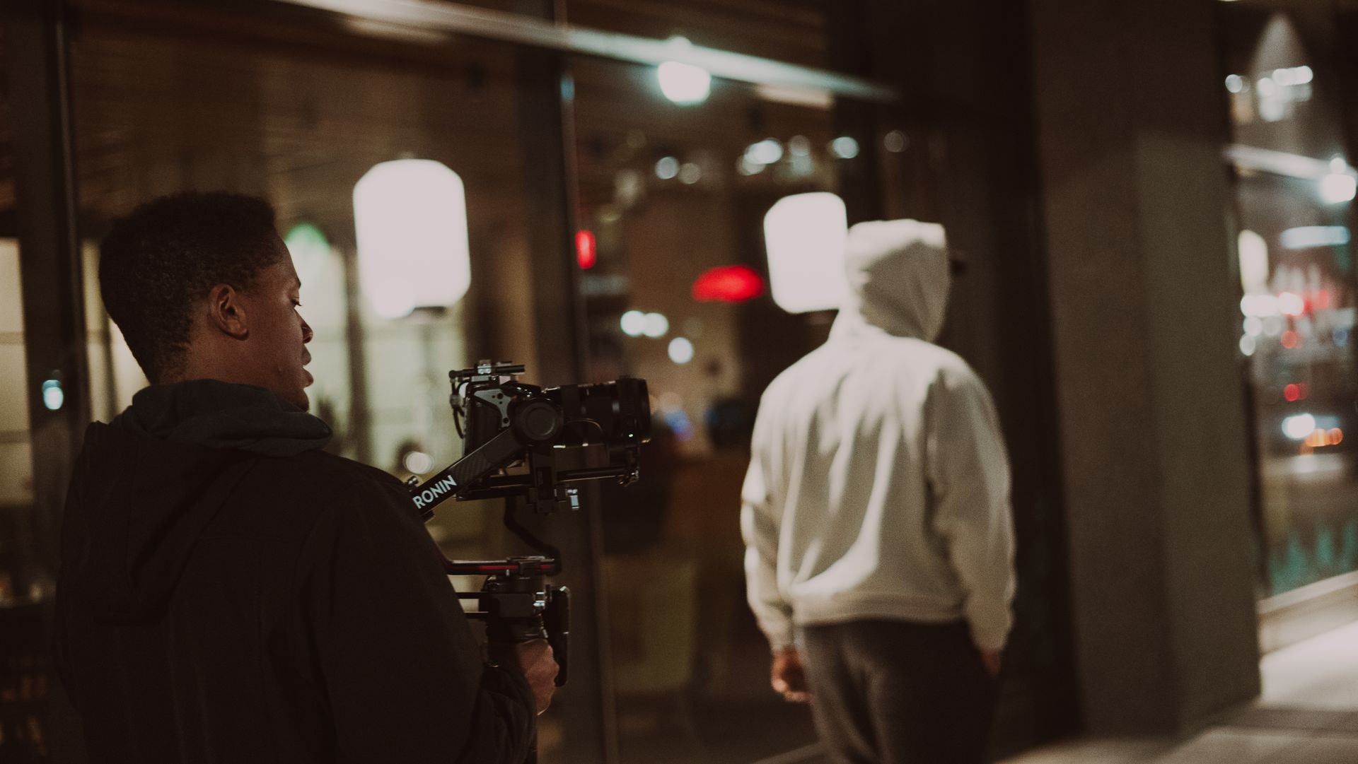 Person filming another individual in a hoodie on a dimly lit street at night.